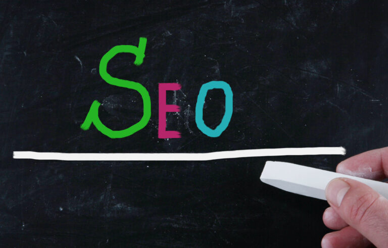 A hand holding a white chalk piece draws a line under colorful &quot;SEO&quot; letters on a blackboard, illustrating the foundation of effective SEO services.