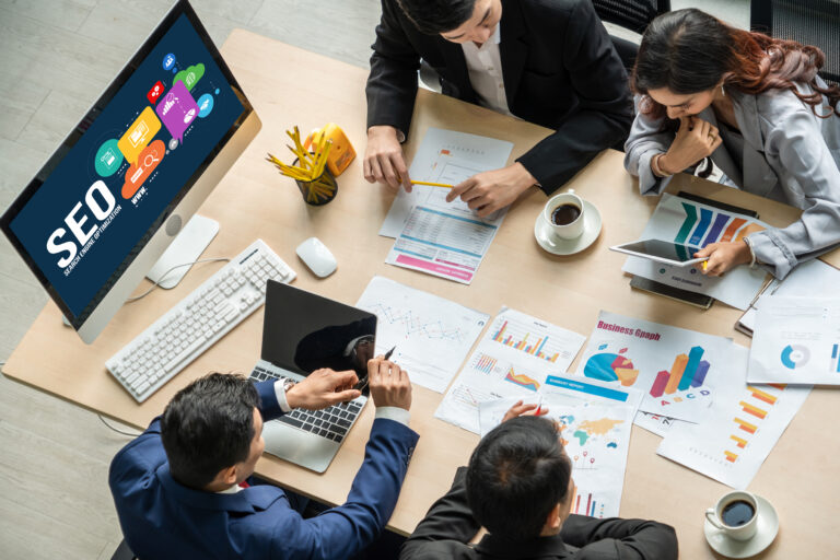 Business meeting with four people discussing charts and graphs at a desk. A monitor displays &quot;SEO&quot; with related icons, highlighting intensive one-day SEO training. Coffee cups and office supplies are scattered across the table, fueling a day of insightful learning.